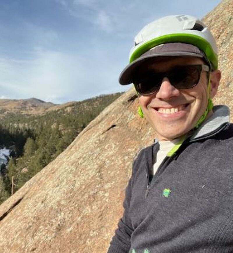 The image shows a man wearing a climbing helmet and sunglasses, smiling at the camera. He is outdoors, possibly rock climbing, as there is a large rock face behind him. In the background, there's a landscape with trees and mountains under a partly cloudy sky. The man is wearing a dark-colored sweater or jacket.